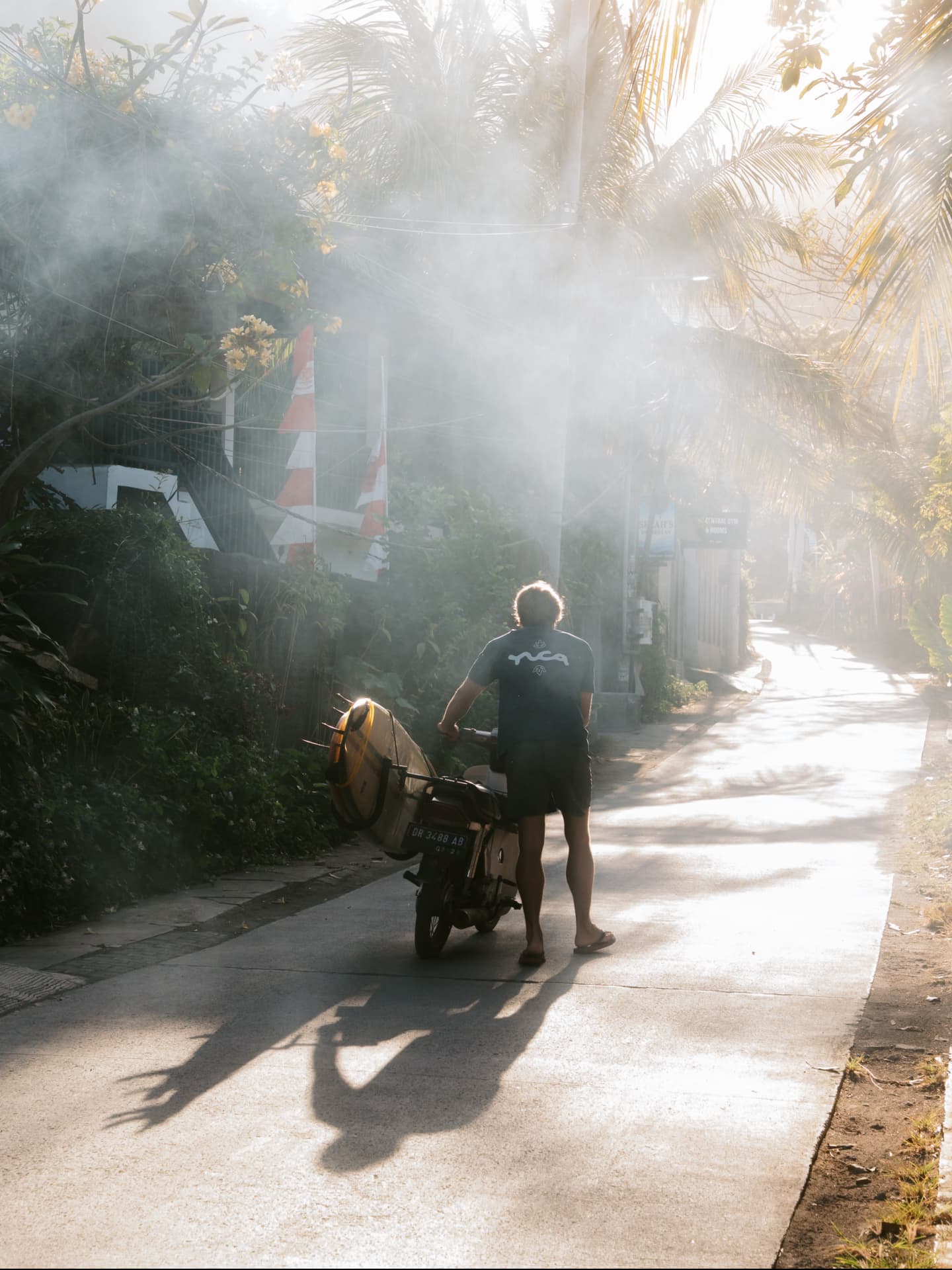 Surfer walking a motorbike down a misty tropical road