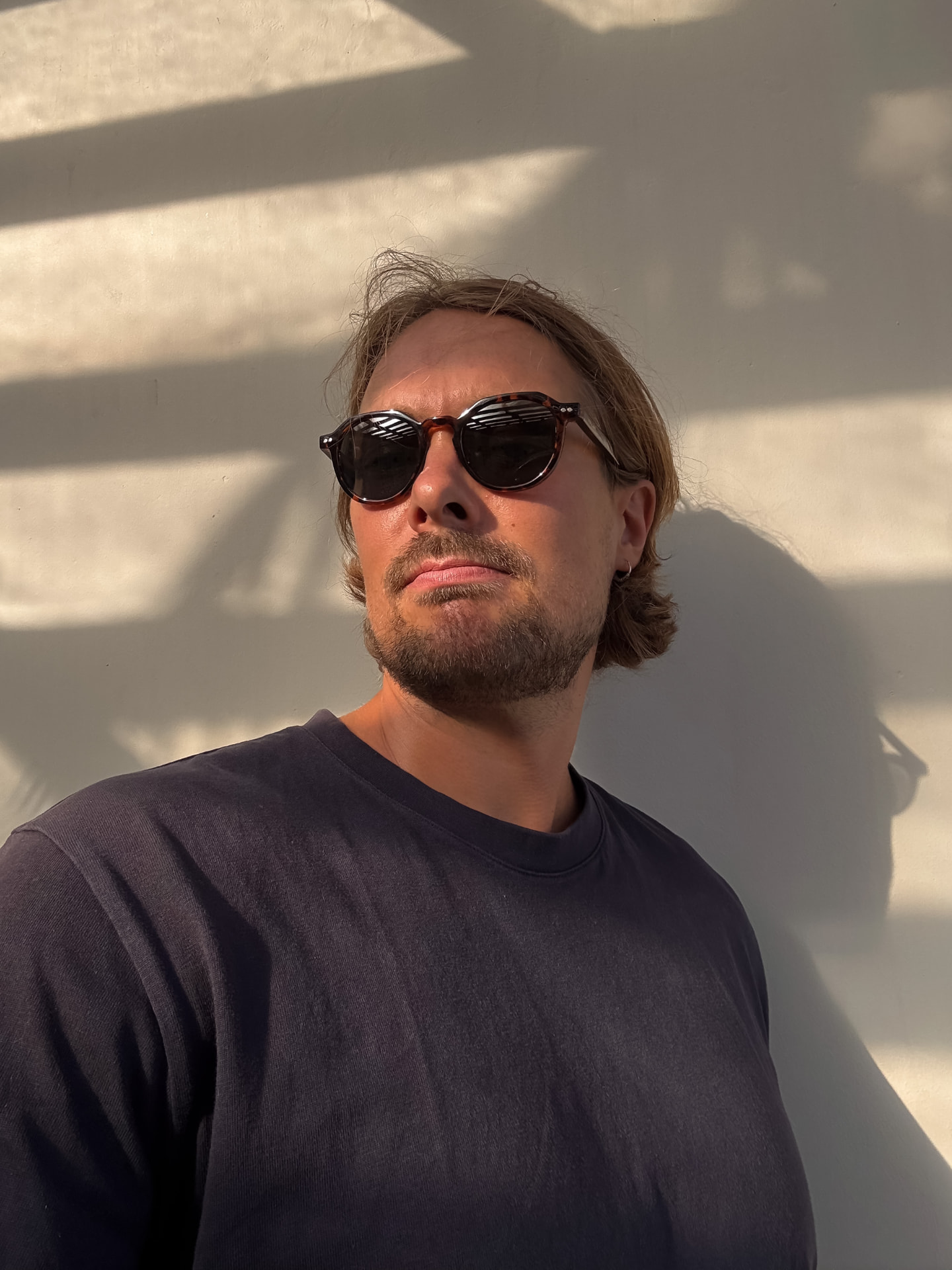 Sunlit portrait of Teemu wearing sunglasses against a white wall